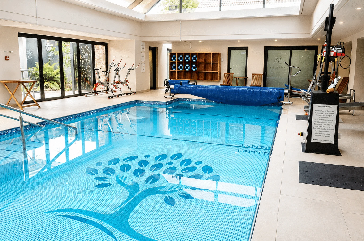 Indoor hydrotherapy pool under a skylight, with a tree mosaic on the pool floor and a hoist beside it